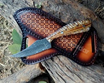 May include: A handcrafted hunting knife with a patterned blade and a handle made of bone and wood. The knife rests in a custom-made leather sheath with a geometric design. The sheath is brown with darker edges.