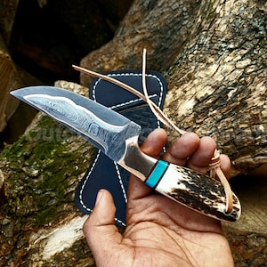 Peut inclure: Un couteau de chasse artisanal avec une lame à motifs et un manche avec un accent turquoise. Le manche est en os et en bois. Le couteau est présenté avec un étui en cuir noir et une lanière en cuir.