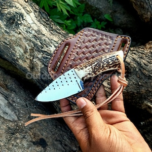 Può includere: Un coltello da caccia con una lama a motivi e un manico in materiale simile all'osso. Il coltello è in un fodero di cuoio marrone con un design intrecciato. Un cinturino in pelle è attaccato al fodero.