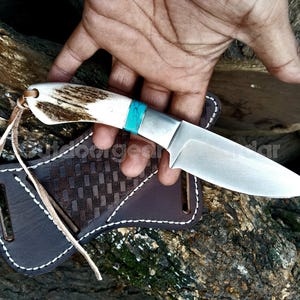 Puede incluir: Un cuchillo de caza artesanal con una hoja de acero pulido y un mango de hueso y detalles turquesa. El cuchillo se muestra con una funda de cuero marrón con un patrón tejido y costuras blancas.