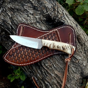 May include: A handcrafted knife with a patterned blade and a bone handle rests in a brown leather sheath. The sheath features a woven design and is secured with a leather strap. The knife and sheath are displayed on a weathered log.