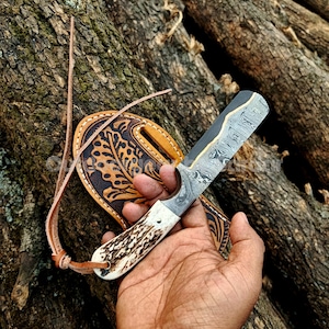 May include: A Damascus steel knife with a bone handle and a leather sheath. The knife blade has a unique pattern. The sheath is brown with a decorative leaf design. The knife is held in a person's hand.