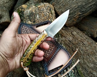 May include: A handcrafted knife with a silver blade and a handle featuring green and yellow accents. The knife is held in a hand, with a brown leather sheath with a geometric pattern visible in the background. A leather strap is attached to the sheath.