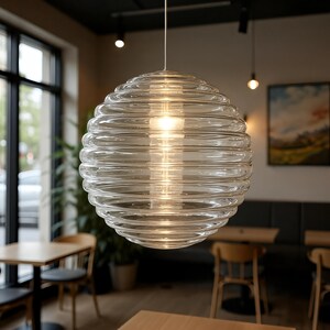 Könnte beinhalten: Eine transparente, kugelförmige Pendelleuchte mit geripptem Design. Die Glühbirne ist im Inneren des Glaskolbens sichtbar. Die Leuchte hängt an einem weißen Kabel in einem Café.