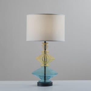 May include: A decorative table lamp with a cream-colored lampshade. The lamp base features a layered design with yellow and blue glass elements. The base sits on a black circular base.