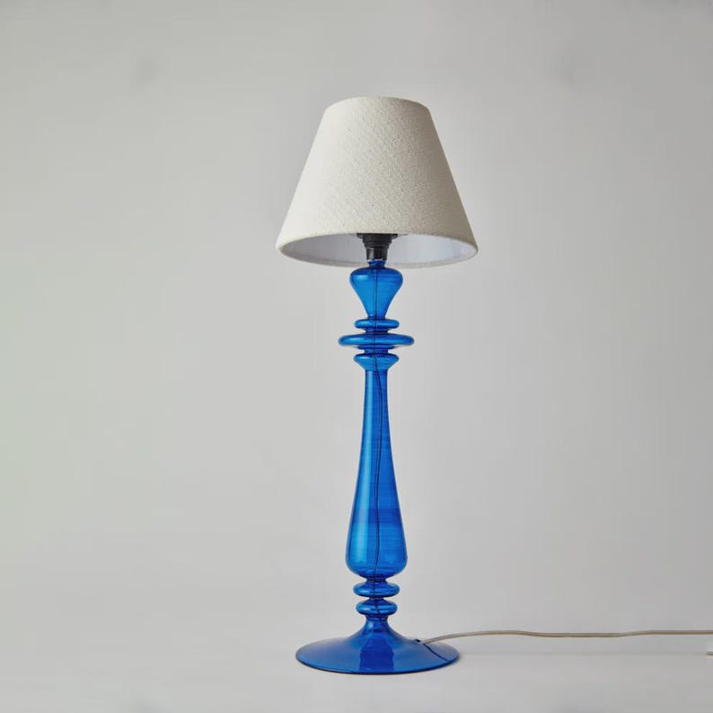 May include: A tall, elegant table lamp with a vibrant blue glass base and a cream-colored lampshade. The base features a slender, elongated design with decorative elements. The lampshade is a classic conical shape, providing soft, diffused lighting.