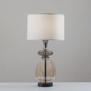 May include: A decorative table lamp with a beige lampshade and a clear, amber-colored glass base. The lamp has a silver-colored metal stem and a black base. The lamp is designed for indoor use and provides ambient lighting.