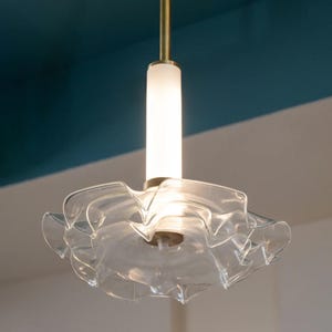 Op de afbeelding: Een hanglamp van helder glas met een gerimpeld ontwerp. De lamp hangt aan een gouden stang en heeft een witte, matte glazen cilinder.