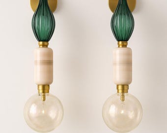 Puede incluir: Dos apliques de pared con un diseño moderno. Cada aplique presenta una parte superior de vidrio verde, una sección media cilíndrica de color crema y una bombilla redonda transparente. Los apliques tienen detalles metálicos dorados y están montados sobre una base circular dorada.