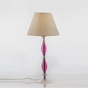 May include: A decorative table lamp with a beige lampshade and a clear base. The lamp's stem features a vibrant pink, oval-shaped design, adding a pop of color. The lamp is designed for indoor use and has a power cord.