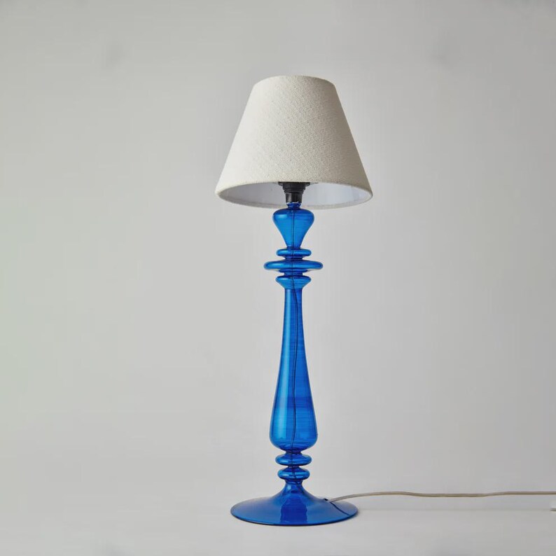 May include: A tall, elegant table lamp with a vibrant blue glass base and a cream-colored lampshade. The base features a slender, elongated design with decorative elements. The lampshade is a classic conical shape, providing soft, diffused lighting.