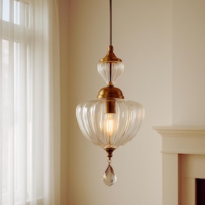 Puede incluir: Una lámpara colgante de cristal transparente y latón. La lámpara tiene una pantalla de cristal acanalado y un cristal decorativo en la parte inferior. El accesorio está suspendido de un cable marrón y está iluminado con una luz cálida.