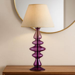 May include: A table lamp featuring a translucent purple glass base with a stacked, bulbous, organic shape. The lamp is topped with a tapered, off-white fabric lampshade that provides a soft, diffused light, creating a balanced and elegant silhouette.