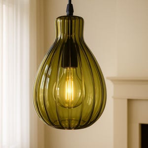 Puede incluir: Lámpara colgante de vidrio verde oliva con diseño acanalado. La lámpara tiene forma de lágrima y un cable negro. Se ve una bombilla cálida dentro de la pantalla de vidrio. El fondo incluye una cortina blanca y una chimenea.