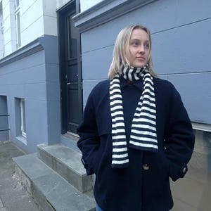 Könnte beinhalten: Eine Frau trägt einen dunkelblauen Mantel mit einem schwarz-weiß gestreiften Schal. Sie steht vor einem Gebäude mit einer grauen Wand.