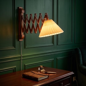 May include: A wall-mounted accordion lamp with a dark wood scissor arm and a pleated white conical shade. The lamp is illuminated, casting a warm glow. A brown leather-bound notebook, a fountain pen, and an inkwell sit on the desk below.
