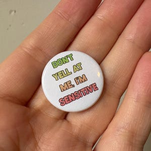 May include: A white button with the text "DON'T YELL AT ME, I'M SENSITIVE" in green, yellow, orange, and red. The button is round and is held in a hand.