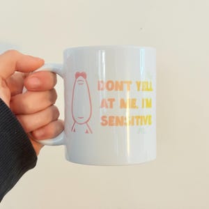 May include: White ceramic mug with a handle, featuring the text "DON'T YELL AT ME, I'M SENSITIVE" in gradient orange and yellow. A cartoon figure is also printed on the mug. The mug is held by a person.