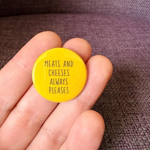 May include: A circular yellow pin-back button featuring the black text MEATS AND CHEESES ALWAYS PLEASES printed in a simple sans-serif font. The button has a smooth, glossy finish and a standard round shape, displaying the phrase clearly on its surface.
