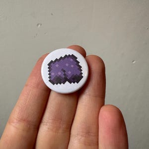 Könnte beinhalten: Ein weißer runder Ansteckbutton mit einem pixeligen lila Design. Das Design stellt eine stilisierte Shorts dar. Der Button wird in der Hand einer Person gehalten. Der Hintergrund ist eine schlichte, hellgraue Wand.