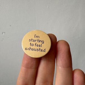 May include: A round, light tan button with the text "I'm starting to feel exhausted" in black. The button is held between two fingers against a neutral background.