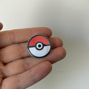 May include: A circular badge with a red and white design, a black outline, and a black circle in the centre. The badge is held in a hand against a white backdrop. The design of the badge resembles a Pokeball.