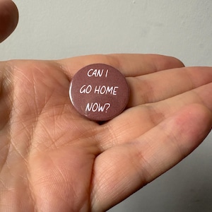 Op de afbeelding: Een ronde, bruinrode button met de witte tekst "CAN I GO HOME NOW?" wordt in een hand gehouden. De button is een eenvoudig accessoire, waarschijnlijk een speld, met een directe boodschap.
