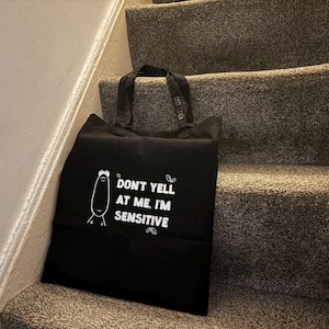 May include: Black tote bag with white text that reads "DON'T YELL AT ME, I'M SENSITIVE." A cartoon character is on the left side of the text. The bag is sitting on a set of stairs with gray carpet.