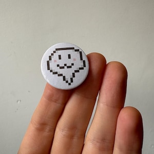 Könnte beinhalten: Ein runder, weißer Knopf mit einem pixeligen Geisterdesign. Der Geist hat einen schwarzen Umriss, schwarze Augen und einen lächelnden Mund. Die Wangen sind rosa. Der Knopf wird in einer Hand gehalten.