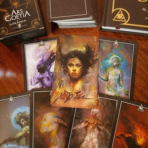 Puede incluir: Un conjunto de cartas del tarot Ars Goetia y un libro. Las cartas presentan ilustraciones detalladas de figuras con temas oscuros y místicos. Los dorsos de las cartas y las cubiertas de los libros son de color marrón oscuro con letras doradas y diseños geométricos.