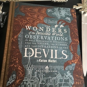 Może przedstawiać: Książka zatytułowana "The Wonders of the Invisible World. Observations As Well Historical As Theological, Upon The Nature, The Number, And The Operations Of The Devils. By Cotton Mather." Okładka książki przedstawia ciemnoniebieskie tło z czerwono-pomarańczowym wzorem smoka. Tytuł książki jest w białym kolorze. Okładka książki zawiera również czarno-białą ilustrację kościoła i człowieka z laską stojącego obok krowy.