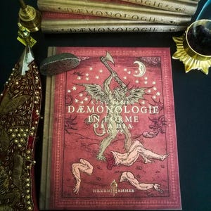 Op de afbeelding: Een rood hardback boek met een gouden titel, "KING JAMES DEMONOLOGIE IN FORME OF A DIA LOGVE", en een gedetailleerde illustratie van een demon die een bijl vasthoudt en een wezen met afgehakte ledematen. Het boek is getiteld "HEXEN HAMMER" in gouden letters.