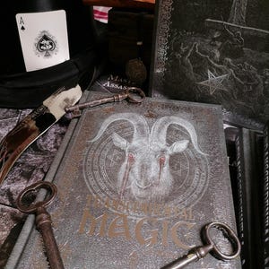 May include: A silver-grey book with the title "The Practical Art of Magic" and a goat with red eyes on the cover. Two antique keys are on top of the book.