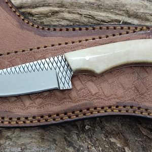 May include: A small, silver knife with a white bone handle and a leather sheath. The knife has a serrated edge and a decorative pattern on the blade.