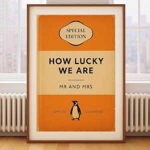 Puede incluir: Impresión enmarcada con un diseño de portada de libro. El título dice "HOW LUCKY WE ARE" con "MR AND MRS" debajo. Hay una ilustración de un pingüino, junto con las palabras "SPECIAL EDITION", "COMPLETE" y "UNABRIDGED".