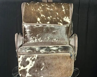 May include: A brown and white cowhide backpack with a top handle and two side pockets. The backpack has a flap closure and a front zipped pocket. The bag is made of leather and has a dark brown trim.