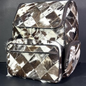 Könnte beinhalten: Ein Patchwork-Rucksack mit einzigartigem Design. Die Tasche besteht aus braunen, weißen und schwarzen Kuhfell-Patches, die in einem geometrischen Muster angeordnet sind. Er hat ein Hauptfach, eine Vordertasche und eine Seitentasche, alle mit Reißverschlüssen. Der Rucksack hat eine dunkelbraune Umrandung.