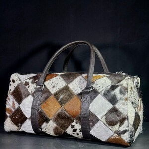 May include: A patchwork duffel bag with a geometric pattern of brown, white, and tan cowhide squares. The bag has dark brown leather handles and straps, and a silver zip.