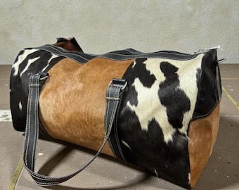 May include: A cowhide duffel bag with a patchwork design. The bag features sections of brown, black and white patterned hide. It has black leather handles and trim, and a zip closure.