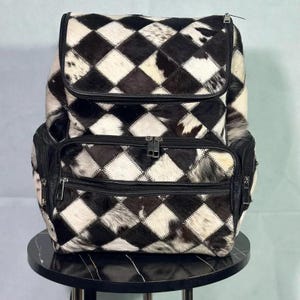 Puede incluir: Mochila de piel de vaca en blanco y negro con diseño de rombos. Cuenta con múltiples compartimentos con cremallera y ribetes de cuero negro. La mochila está sobre una mesa negra.