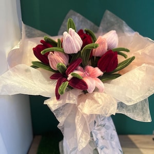 Peut inclure: Un bouquet de fleurs en tissu faites à la main, comprenant des tulipes et des lys, dans des tons de rose et de bordeaux, avec des tiges vertes. Les fleurs sont enveloppées dans du papier de soie et du papier floral décoratif.