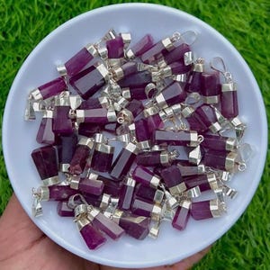May include: A collection of purple gemstone pendants with silver-colored metal caps and loops, displayed in a white bowl. The gemstones have a rectangular shape and are a deep, rich purple color. The pendants are scattered, creating a visually appealing arrangement.