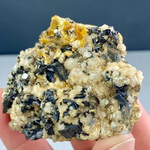 May include: A close-up of a mineral specimen featuring a cluster of black, shiny crystals. The crystals are embedded in a matrix of beige and white minerals, with patches of yellow-orange crystals. The overall texture is rough and uneven.