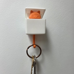 May include: White key holder with a cat face on the front. The cat is orange and has a smiling expression. A metal key ring with keys is hanging from the holder.