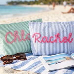 May include: Two seersucker cosmetic bags, one mint green with "Chloe" in pink script, the other blue and white striped with "Rachel" in pink script. Sunglasses and a magazine rest on a striped towel on the beach.