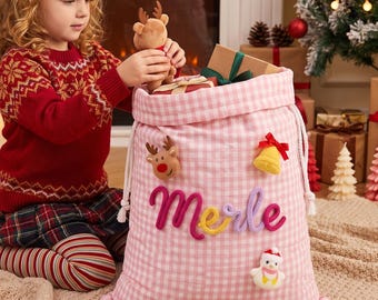 Op de afbeelding: Een lichtroze en witte gingham stoffen cadeautas met de naam Merle in kleurrijke garenletters. De tas is versierd met pluche applicaties van een rendier, een gele bel met een rode strik en een sneeuwpop, en heeft witte trekkoorden.