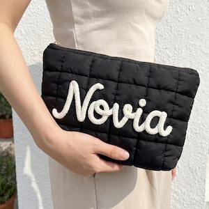 May include: Black quilted clutch with the word "Novia" embroidered in white cursive. The clutch has a rectangular shape with a gold zipper. The background is a neutral color.