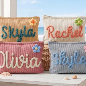 May include: Four rectangular corduroy zip pouches in tan, cream, dusty rose, and light blue. Each pouch features a personalised name in raised, cursive yarn lettering and a small, crocheted flower accent in a contrasting colour.