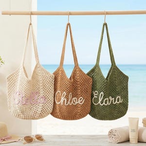 May include: Three woven tote bags in cream, tan, and olive green hang from a wooden rod. Each bag has a name embroidered on it: Stella, Chloe, and Clara. The background shows a beach scene with a hat, sunglasses, and towels.