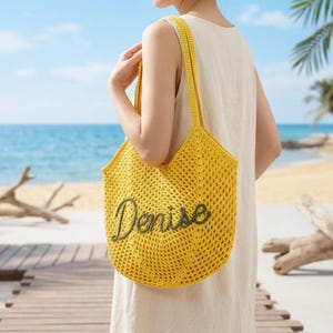 Pu&ograve; includere: Una borsa tote gialla all'uncinetto con il nome "Denise" ricamato in verde. La borsa ha una tracolla ed &egrave; tenuta da una persona. Lo sfondo mostra una scena di spiaggia con una passerella di legno e l'oceano.